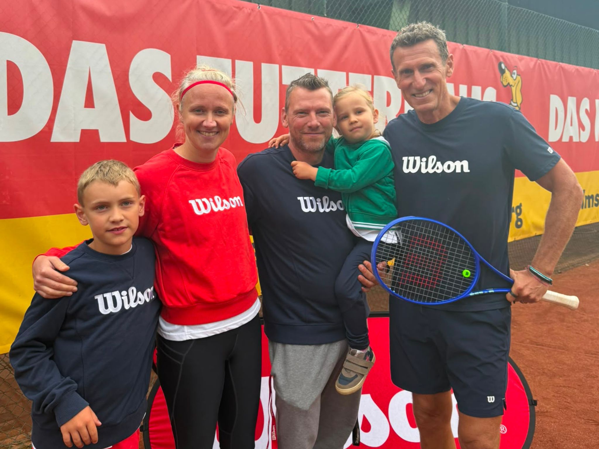Der Cheftrainer des TV Uetersen, Björn Keller, steht mit seiner Familie und Patrick Kühnen auf einem Tennisplatz. Alle lächeln in die Kamera und tragen Sportbekleidung, überwiegend von Wilson. Patrick Kühnen hält einen blauen Tennisschläger. Im Hintergrund ist ein großes Banner des TV Uetersen zu sehen.